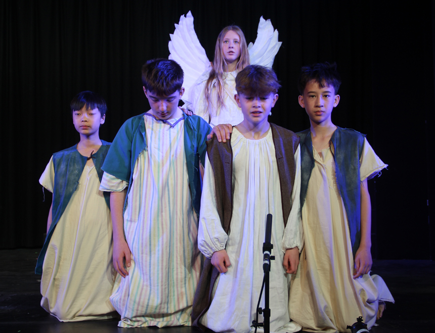 four children kneeling on a stage with a child dressed as an angel behind