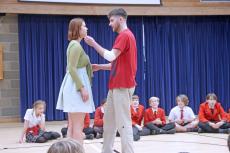 Two actors standing up in front of a group of children seated on the floor watching the actors perform