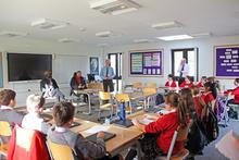 School children sitting at desks in a u-shape facing a group of visitors at the front of the classroom