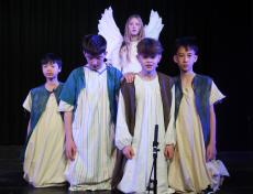 four children kneeling on a stage with a girl behind dressed as an angel