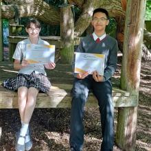 two children sitting on a bench holding up maths certificates