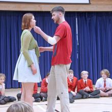 Two actors standing up in front of a group of children seated on the floor watching the actors perform