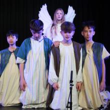 four children kneeling on a stage with a girl behind dressed as an angel